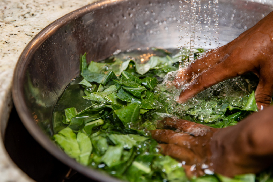 African American hands rinse chopped collard greens in stainless steel bowl under running water.